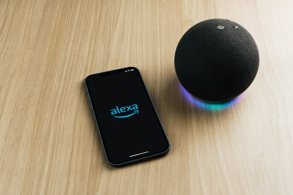 Amazon Alexa smart speaker next to a smartphone with the Alexa app open.