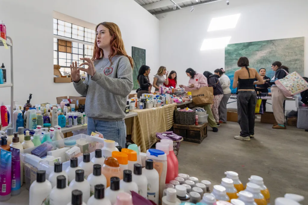 A teenage girl gestures while speaking inside a large room filled with tables covered in hair, skincare and beauty products as other girls and volunteers browse in the background.