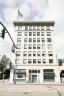 A multi-story white building with large windows and “Altadena Girls” signage on the ground floor sits on a street corner beneath a cloudy sky.
