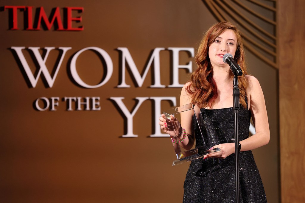 A teenage girl stands at a microphone holding a glass award on a stage with “TIME” branding behind her.