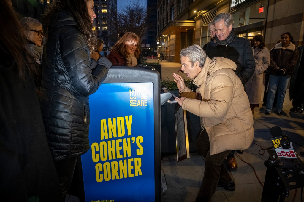 Andy Cohen leaning into a trash can with a sign reading "Andy Cohen's Corner" while people watch and smile.