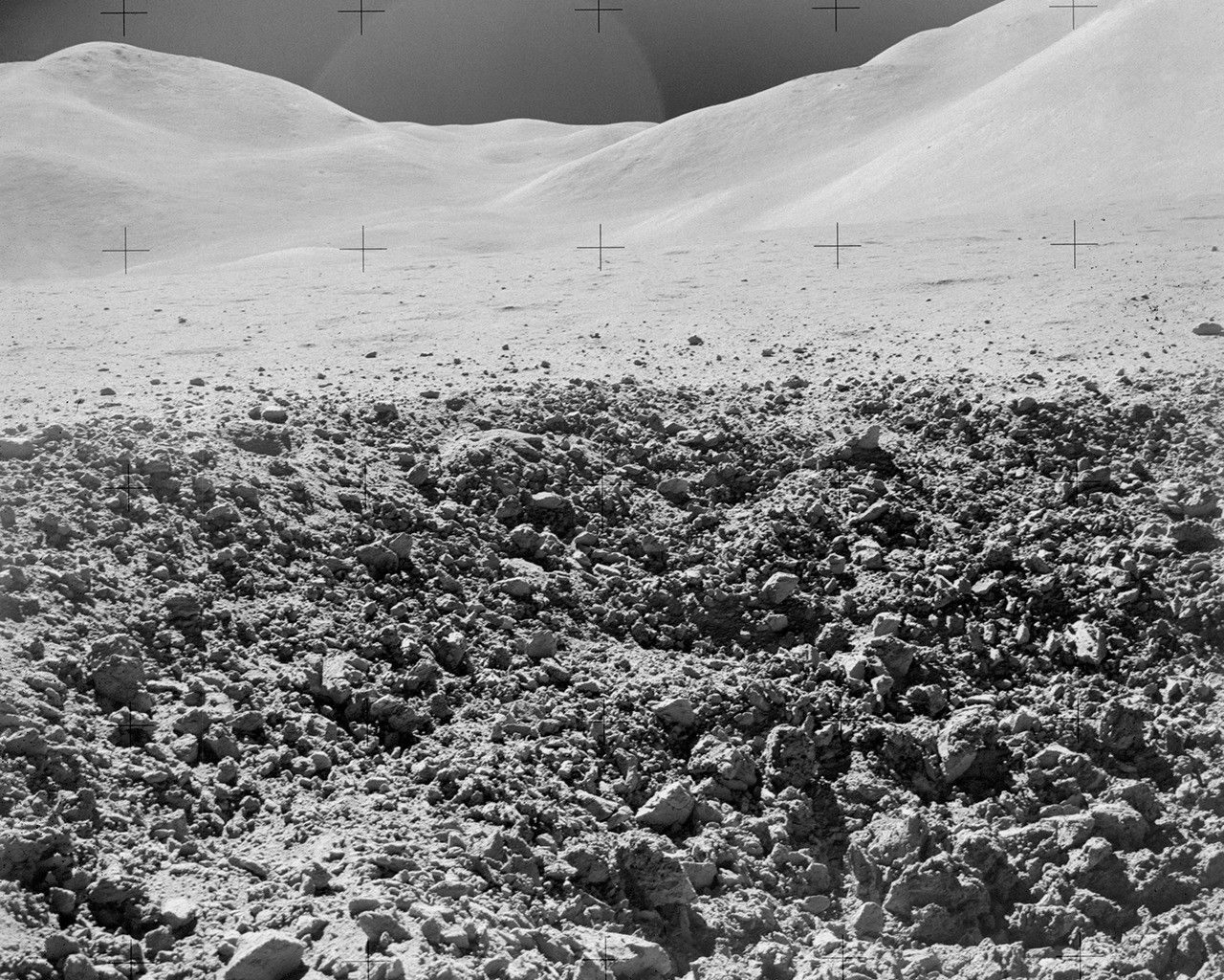 View from surface of lunar crater. The foreground looks like an expanse of rocky rubble. In the background, lighter-colored, dune-shaped hills rise under a dark sky.