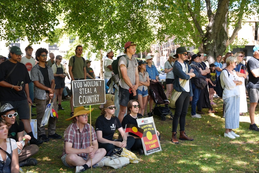 A crowd of people sitting and standing under trees. One holds a sign that says "You wouldn't steal a country".