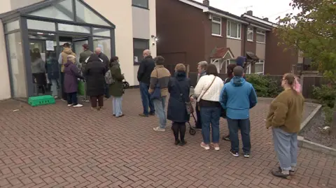 BBC A queue of about a dozen people outside the door of a dental practice, with the queue then carrying on out of frame. People are dressed in coats and jumpers.