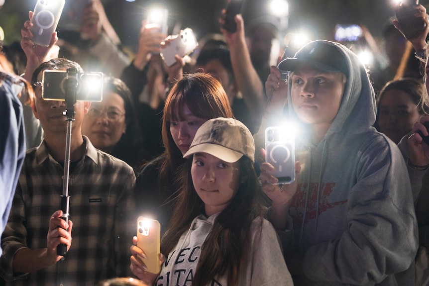 A group of people hold phones with the torches on and lit candles in the night.
