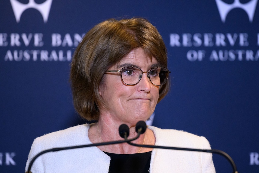 A bespectacled, auburn-haired woman in a light jacket smiles grimly as she speaks at a podium.