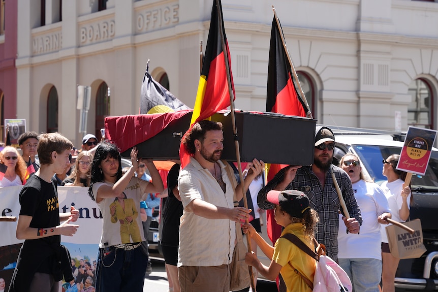 A man takes an Aboriginal flag from a young girl wearing yellow, he holds a coffin, a crowd gathers behind them