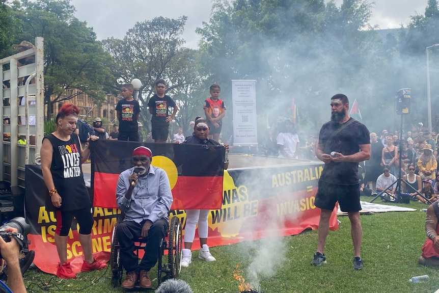 An indigenous elder speaking in front of Indigenous flag at Invasion Day event