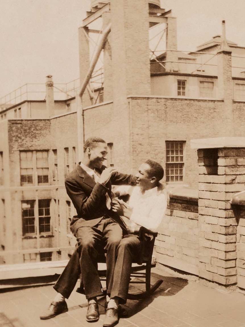 A man sits on another's lap on a chair on a rooftop building.