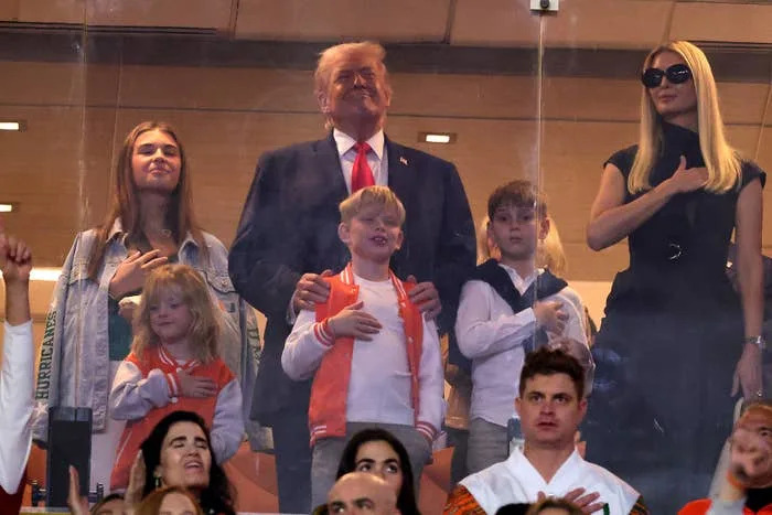 A group of people stands in a stadium box, some with hands on hearts. They appear to be watching an event, showing a moment of attention and respect