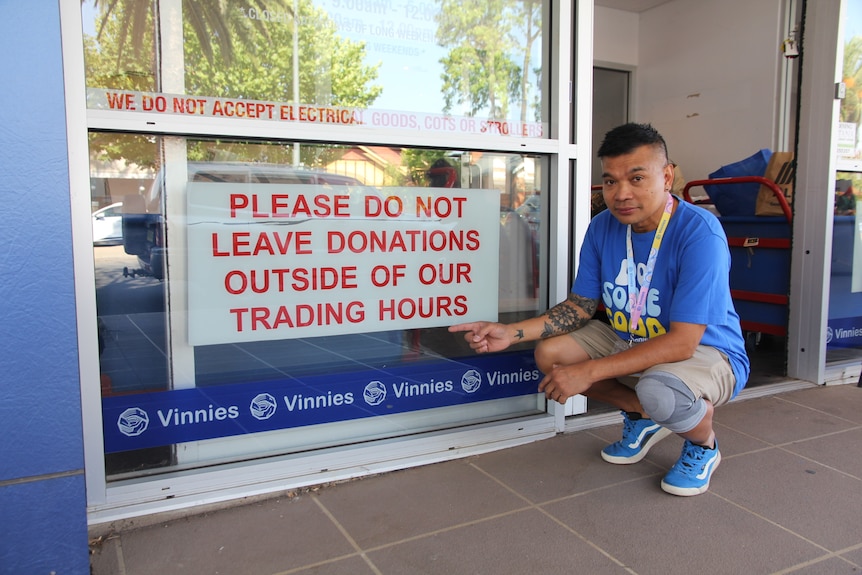 A man in a blue shirt and black hair points to a sign that says not to leave donation items outside their office hours