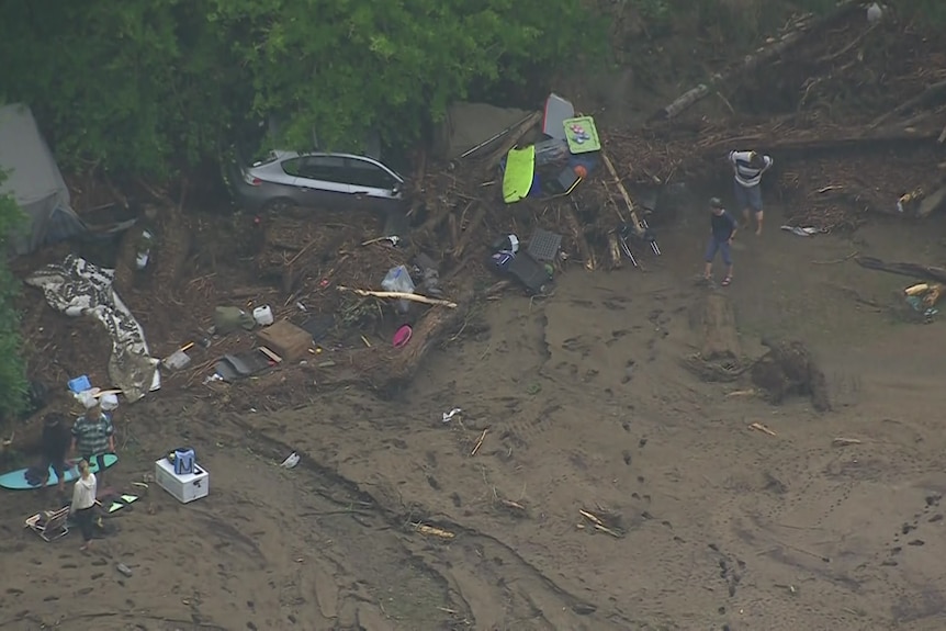A car is half buried in mud under a tree with other objects and debris scattered around.