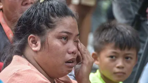 AFP via Getty Images A close up shot of a woman wiping a tear away from her eye at the scene of the landfill site, while a small boy looks across at her.