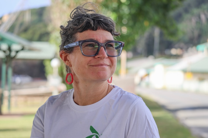 A woman with red earings and glasses wearing a white t-shirt looks away from the camera with a slight smile outdoors.
