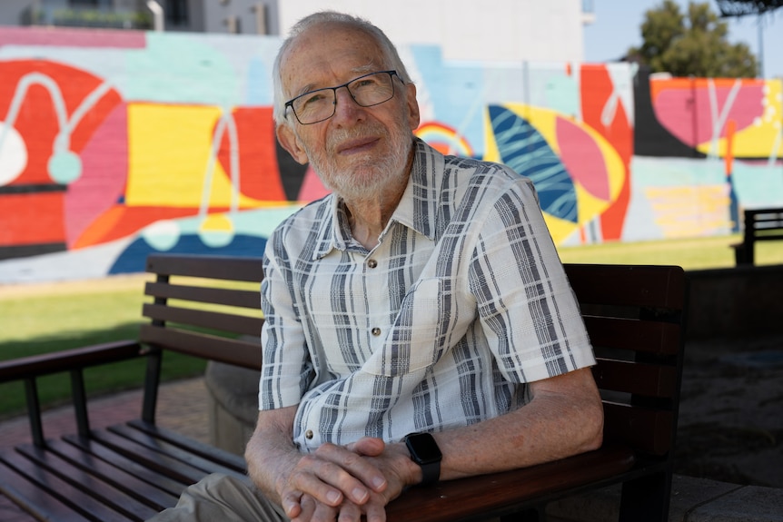 Bruce rests his arm on a park bench while sitting in a park with street art in the background