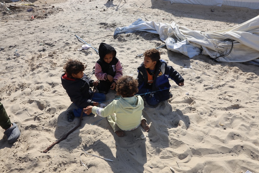 Four young children sitting in a circle on sand, near them is a broken tent tarp.