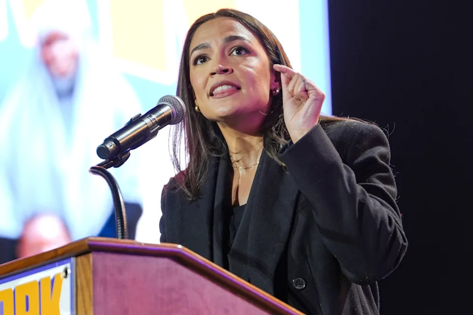 Person speaking passionately at a podium with a microphone, gesturing with hand for emphasis, wearing a dark blazer