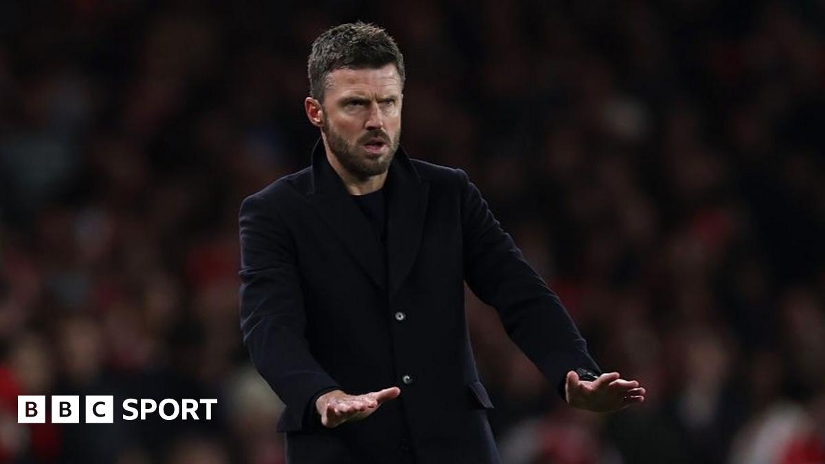 Manchester United interim head coach Michael Carrick makes a palms-down gesture that appears to be calling for calm as he wears a dark coat over a dark top