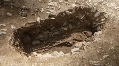 Kevin Church/BBC News A human skeleton is lying in an oval-shaped grave cut into limestone bedrock. The bones are positioned naturally, with the skull at one end and legs extended. The ground surrounding the grave is light brown and cracked.