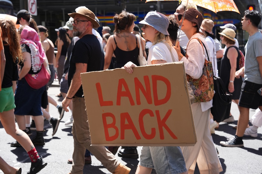 A person walks holding a cardboard sign that says "land back".