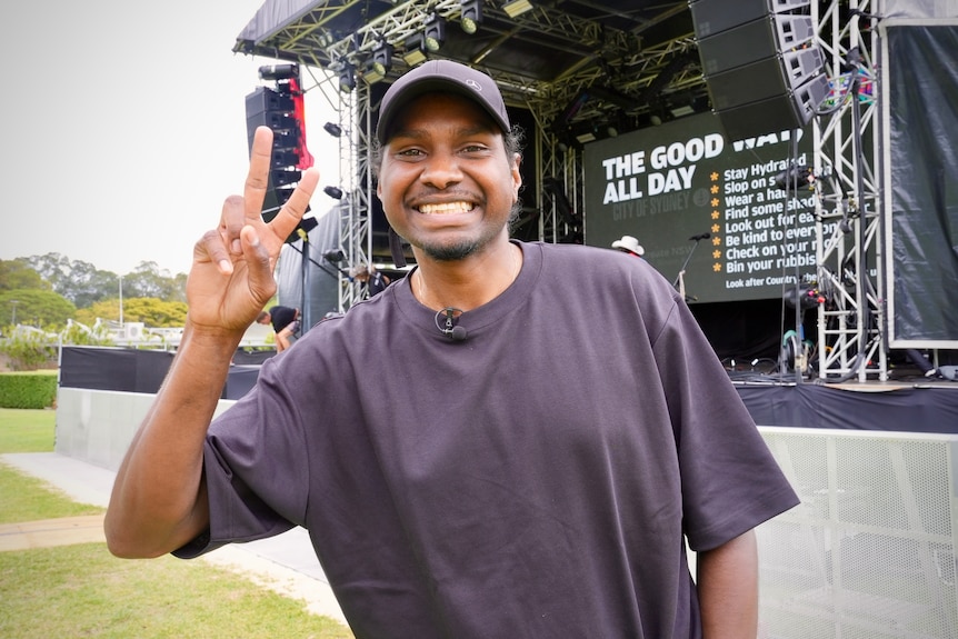 An indigenous man in dark t-shirt and hat smiling at the camera in front of a stage
