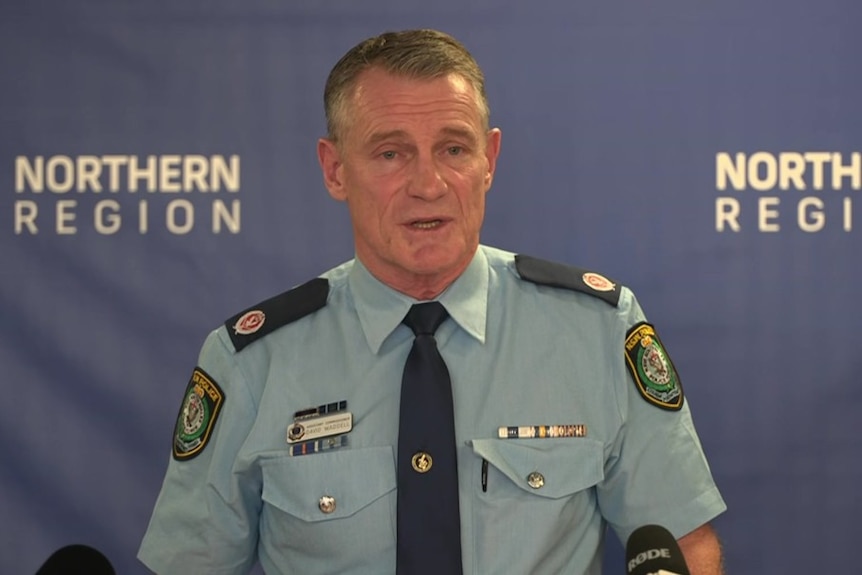 A middle-aged man in a police uniform stands in front of a branded backdrop and speaks to the media.