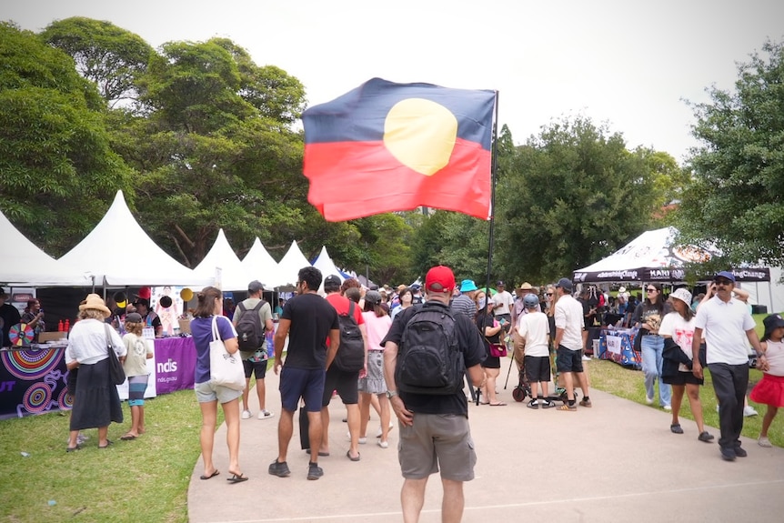 A crowd walking past tents with one person carrying an Aboriginal flag