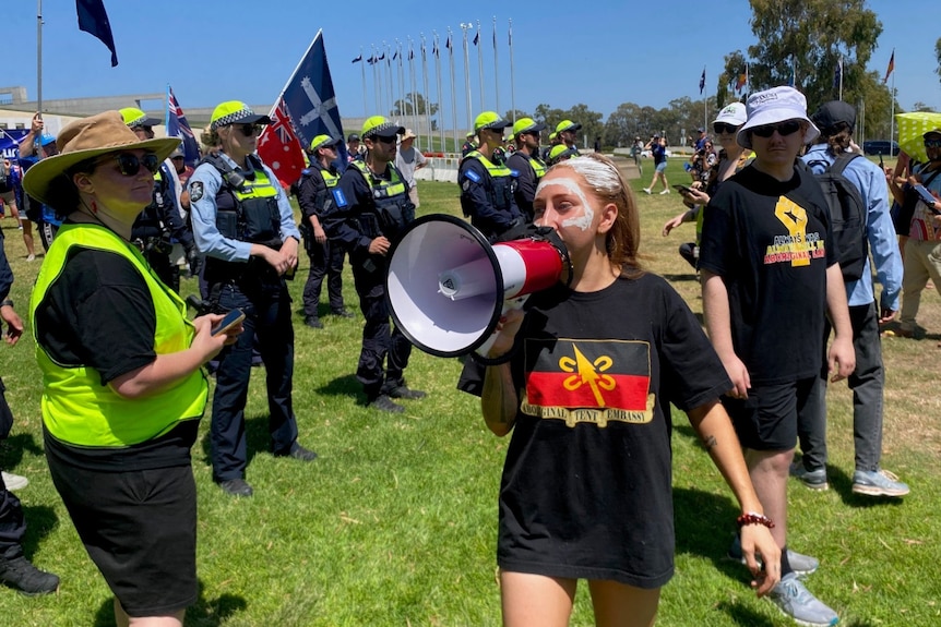 A protestor wearing an Aboriginal Tent Embassy t-shirt shouts into a megaphone while police look on.