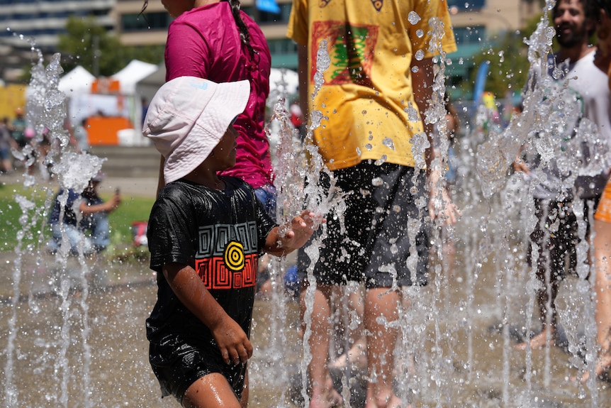 A young child wearing a shirt with the Aboriginal flag on it plays in a water fountain.