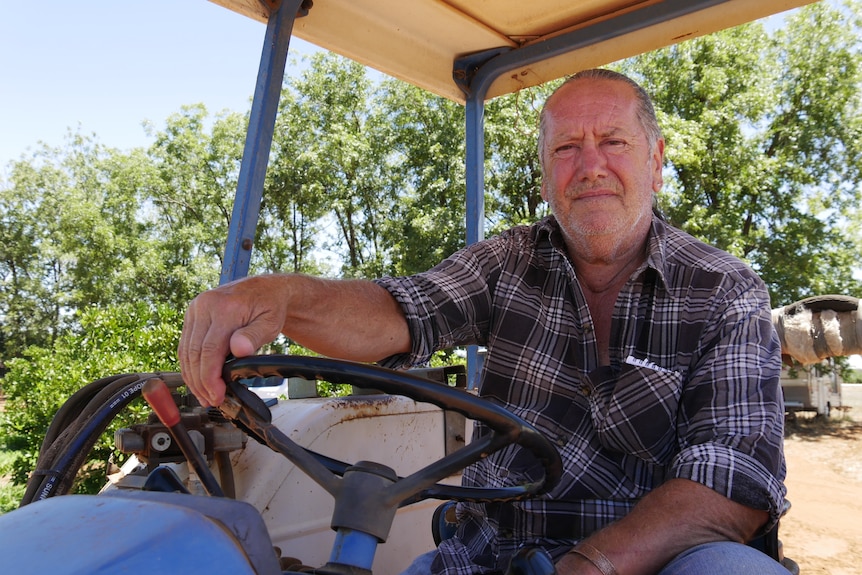 A man with grey hair sits in a tractor holding the wheel wearing a long sleeved flannelette shirt 