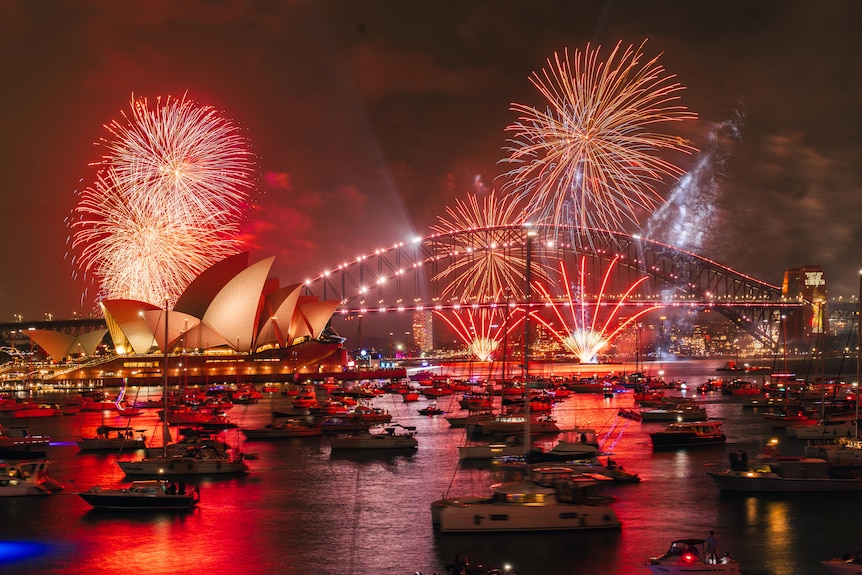 Sydney's harbour with the Opera House and bridge illuminated by red fireworks with boats in the water,