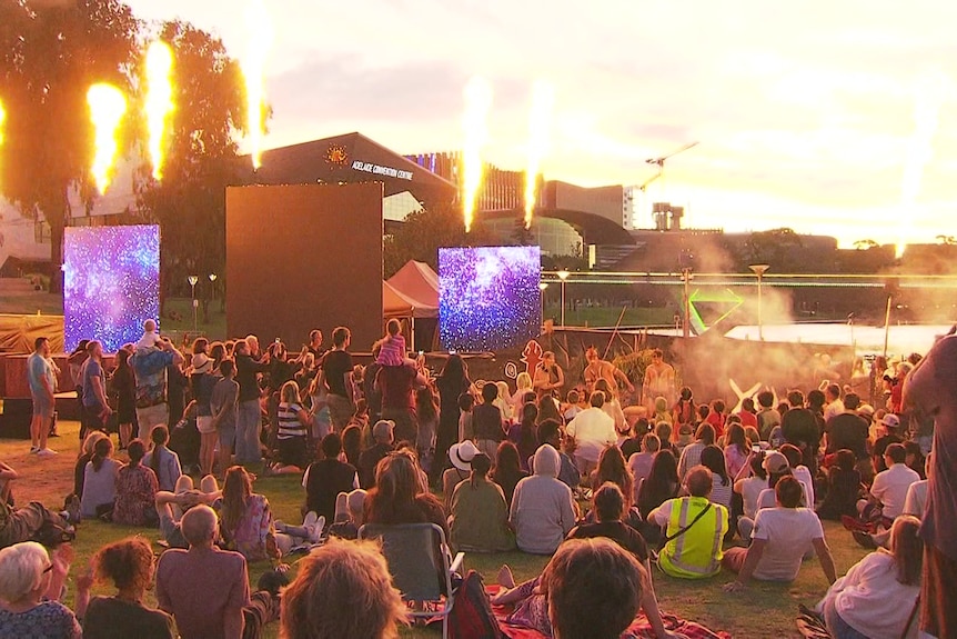 A large seated crowd look at screens with performances and pyrotechnics by a river