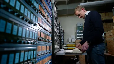 Man in jeans, jumper and a shirt reads an old book that is resting on a steel table in the archives. Tiny boxes that are about the size of printer ink cartridges are neatly stacked on the walls