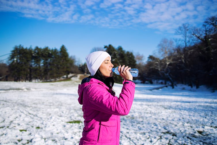 A woman wearing a white hat and a bright pink jacket sips from a water bottle while standing outside on a winter's day.