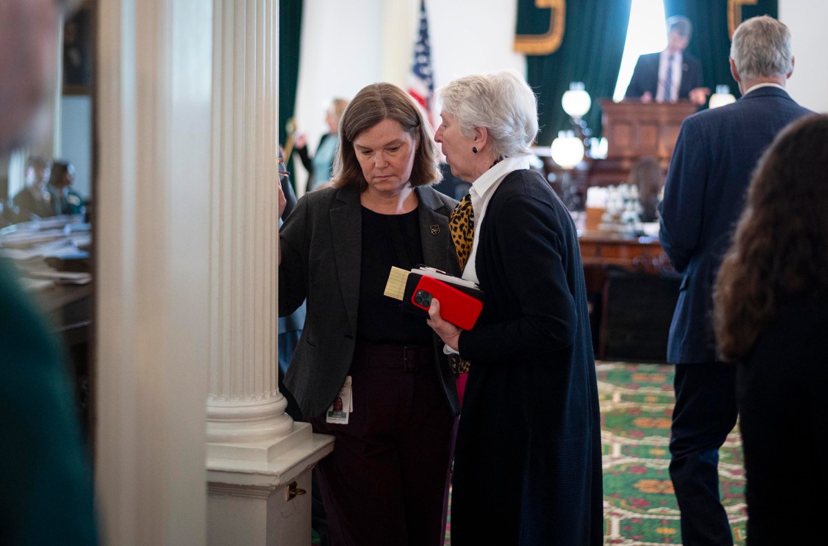 Two women stand near a column in a formal room; one holds a red notebook and speaks to the other, who looks down with a serious expression. Other people are visible in the background.