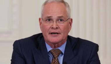 Darren Woods speaks at a meeting, wearing a dark suit, light blue shirt and patterned brown tie.