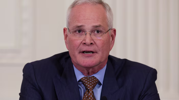 Darren Woods speaks at a meeting, wearing a dark suit, light blue shirt and patterned brown tie.
