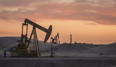 Pumpjacks operate on sandy terrain at sunset with industrial equipment in the background