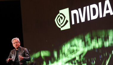 Jensen Huang speaks on stage, gesturing with his hands, with a large Nvidia logo displayed on a screen behind him.