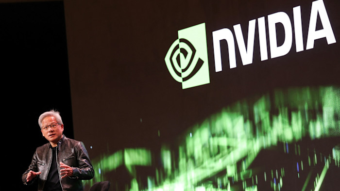 Jensen Huang speaks on stage, gesturing with his hands, with a large Nvidia logo displayed on a screen behind him.