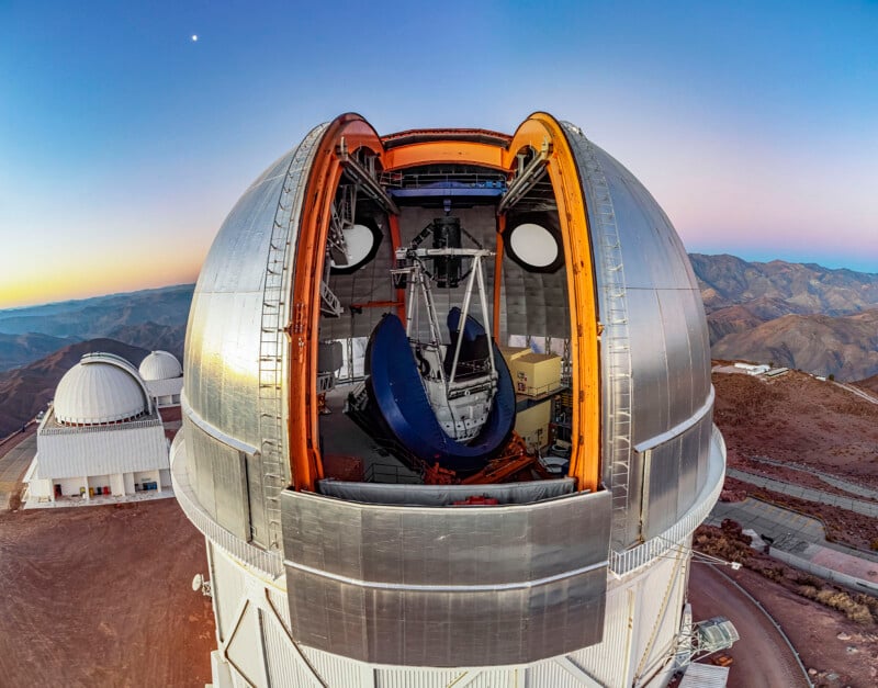 A large astronomical observatory dome with its retractable roof open, revealing a massive telescope inside. Other observatory domes and a rugged, mountainous landscape appear in the background at dusk.