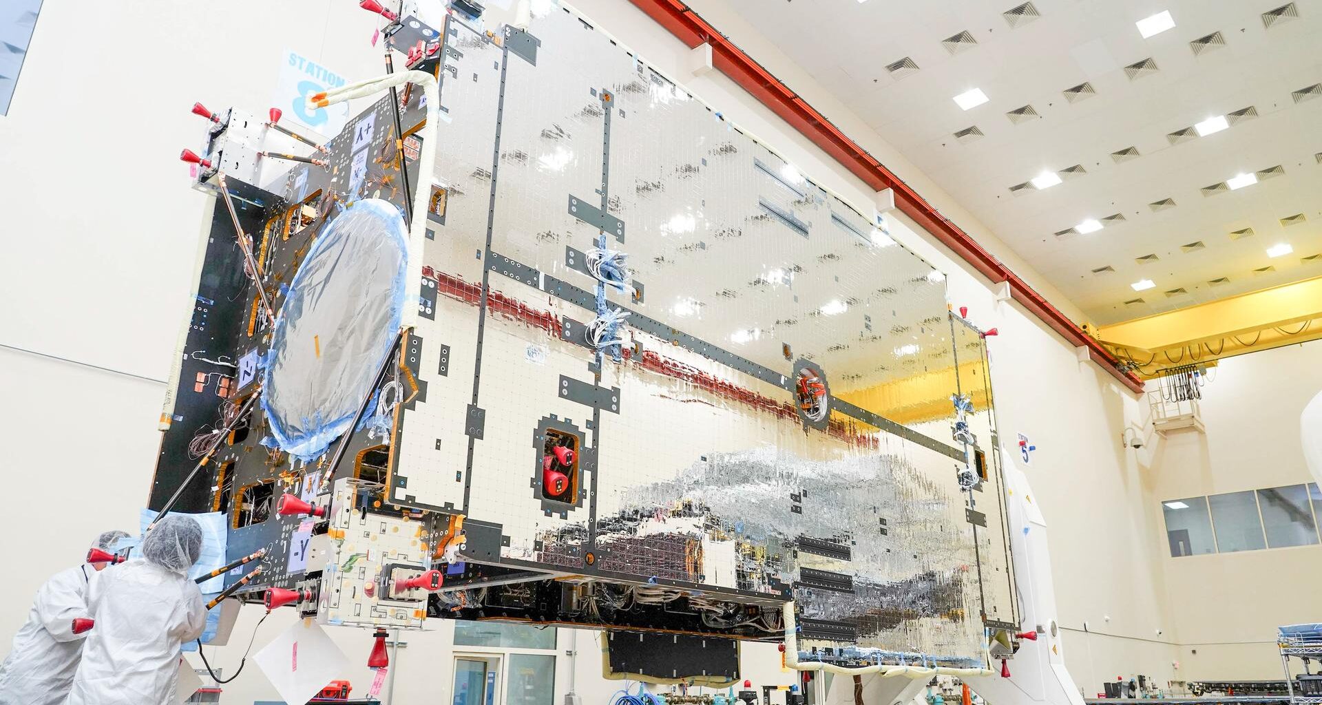 The primary structure of Gateway’s Power and Propulsion Element stands inside a high-bay cleanroom at Lanteris Space Systems in Palo Alto, California. The large rectangular structure is covered in reflective silver-colored panels. Two technicians in white cleanroom suits work near the base of the structure.