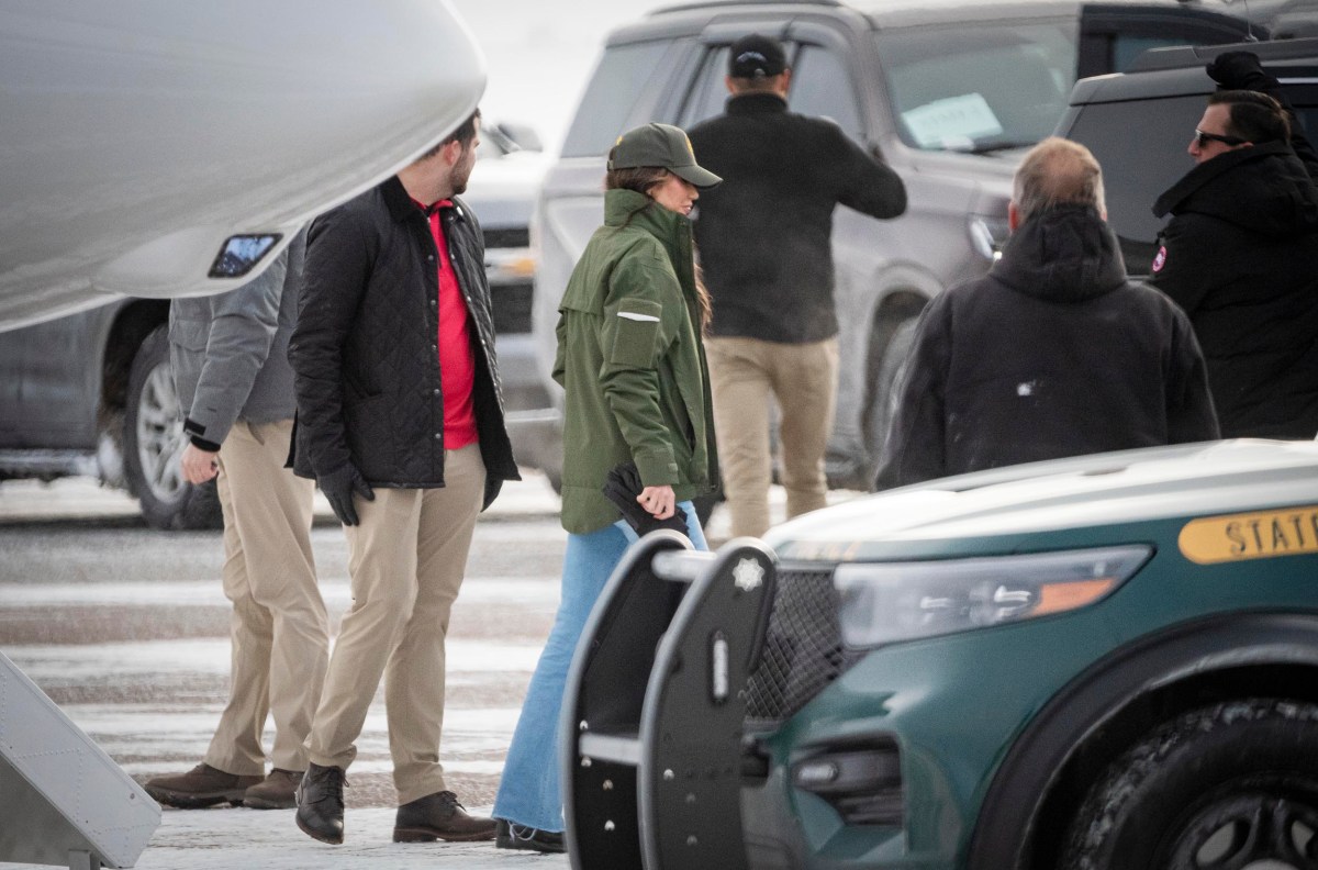 A group of people stands near vehicles and a plane on a snowy tarmac. A woman in a green jacket and cap walks alongside others.
