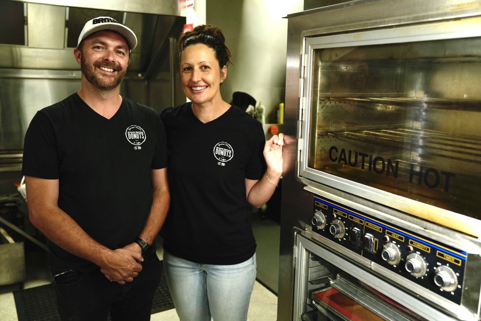 Two people smiling at the camera, stand next to an oven.