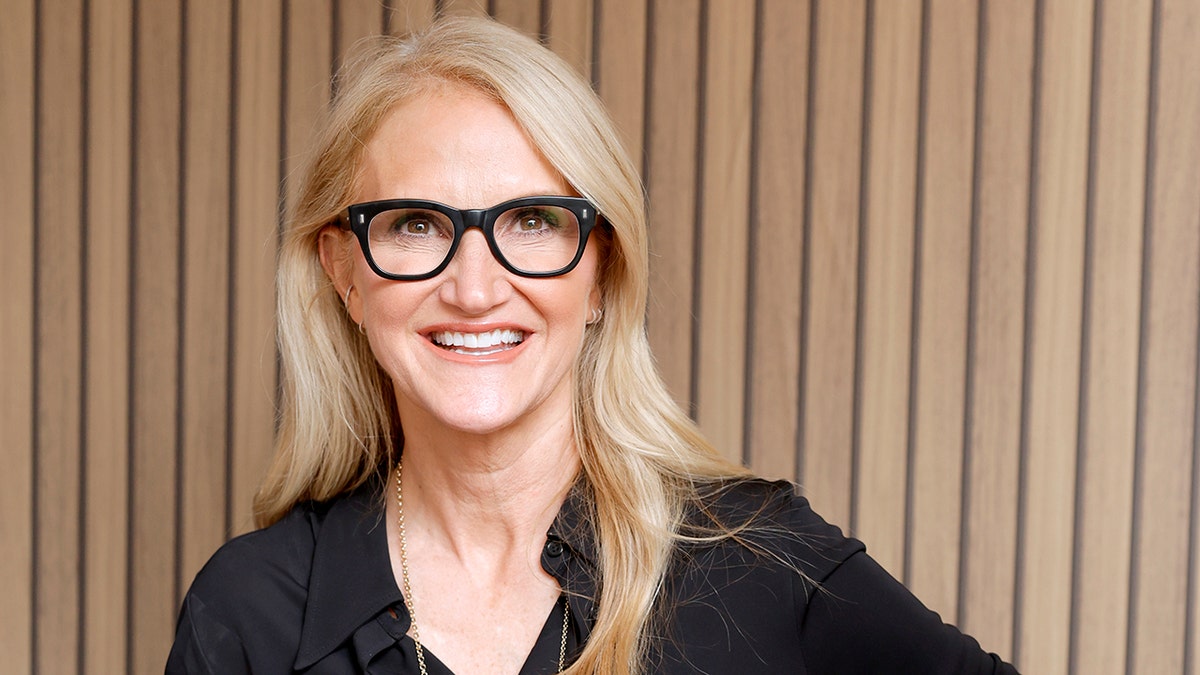 Mel Robbins wearing a black blouse and black framed glasses, in front of a brown paneled background.