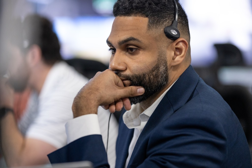 A man with close cropped hair and a short beard, wearing a dark suit and a headset, focuses intently.