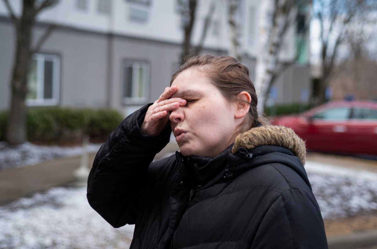 A person stands outdoors, eyes closed, with their hand on their forehead. They wear a black jacket. Snow and buildings are in the background.