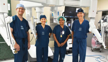 A diverse surgical team stands proudly in an operating room, wearing blue scrubs. They are posed in front of advanced robotic surgical equipment, smiling confidently.