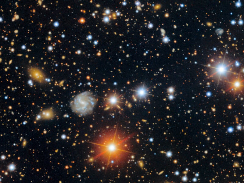 A dense field of stars and distant galaxies scattered across deep black space, with several bright stars, glowing galaxies, and points of multicolored light visible throughout the image.