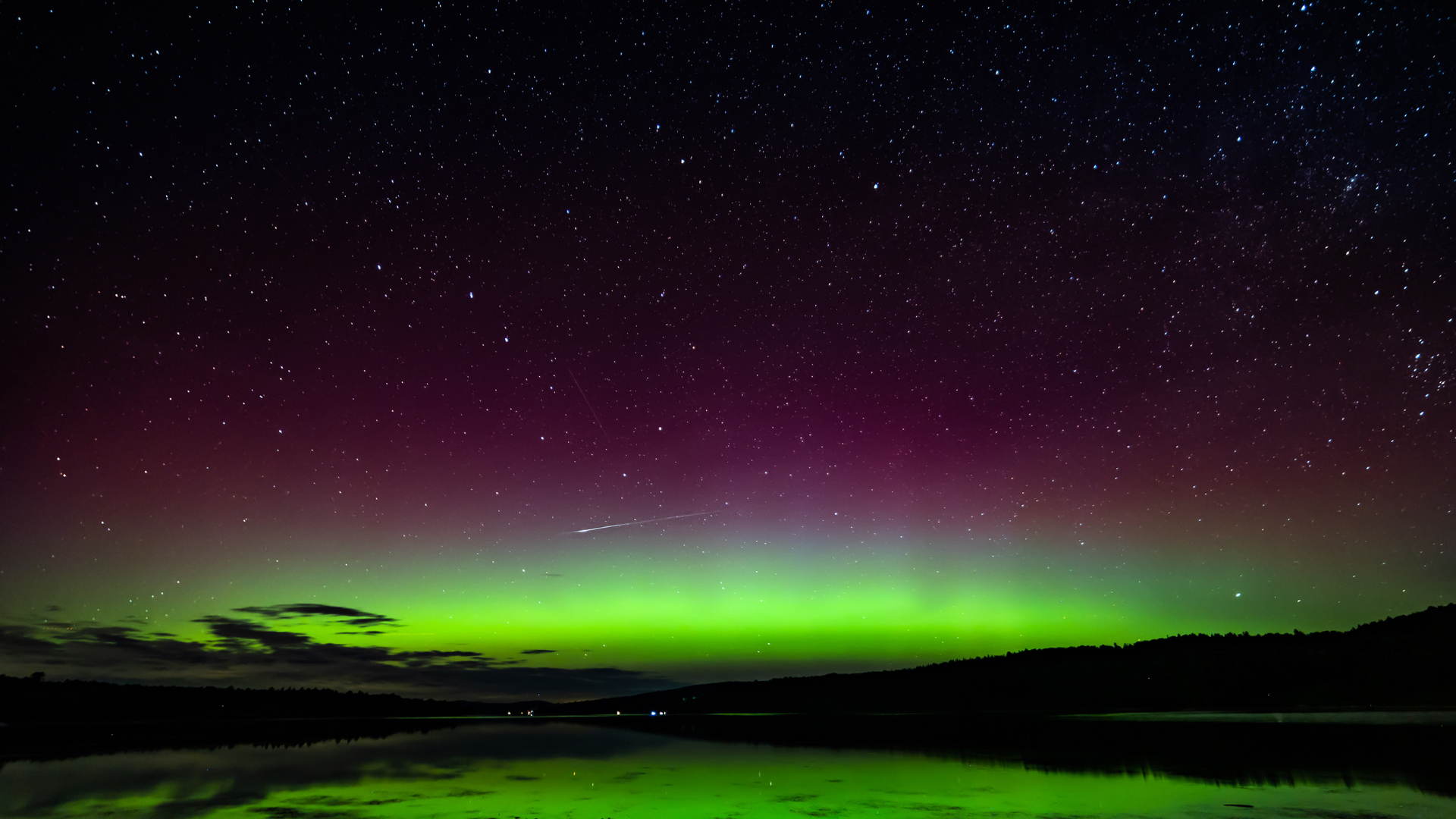 Glowing green and red northern lights are reflected in a still lake with a streak of light toward the left of the image
